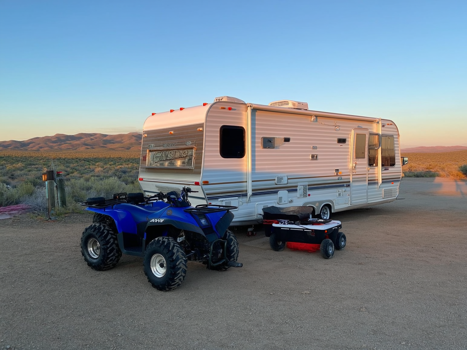 Two ATVs and a small enclosed gear trailer parked and secured beside a full-hookup travel trailer at an RV site near Casper Wyoming, golden hour, sagebrush hills in the background